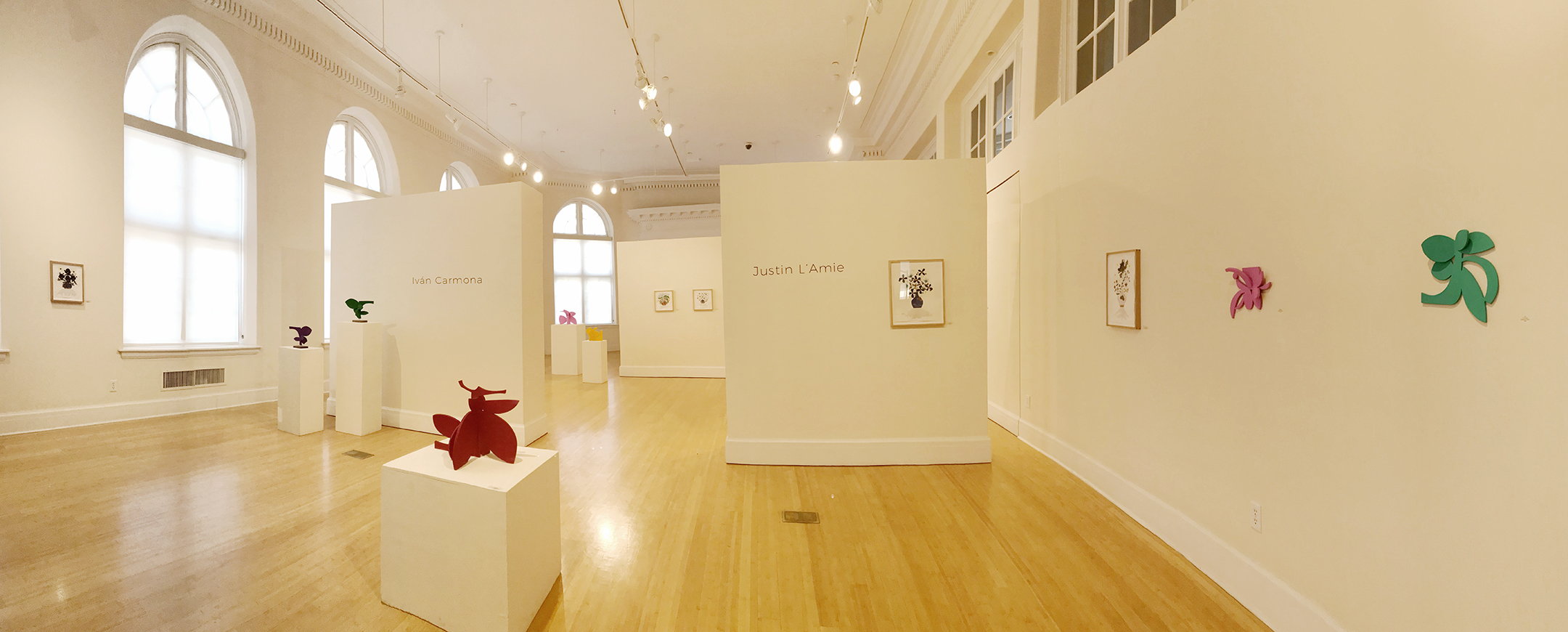 Full view of gallery with large windows and wood floors. with colorful sculptures and paintings on the walls. 