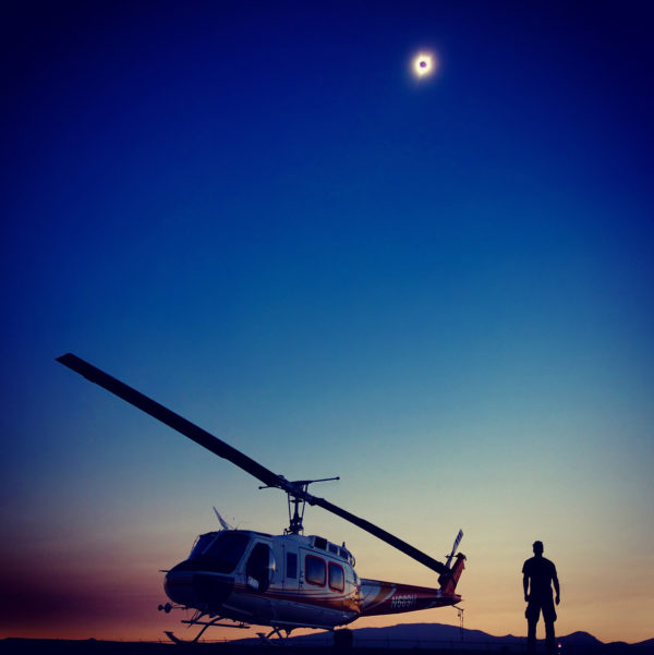 Helicopter and man silhouetted against sky at dusk with lunar eclipse above