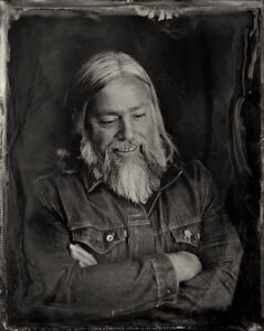 A photo in the style of an old tin type of a man with long gray hair and long beard with arms crossed, wearing a denim jacket.